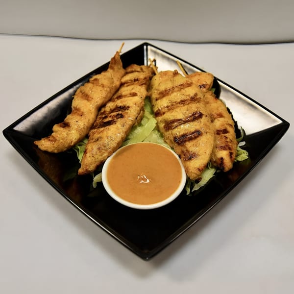 Grilled chicken satay skewers with peanut sauce and lettuce on a black square plate.